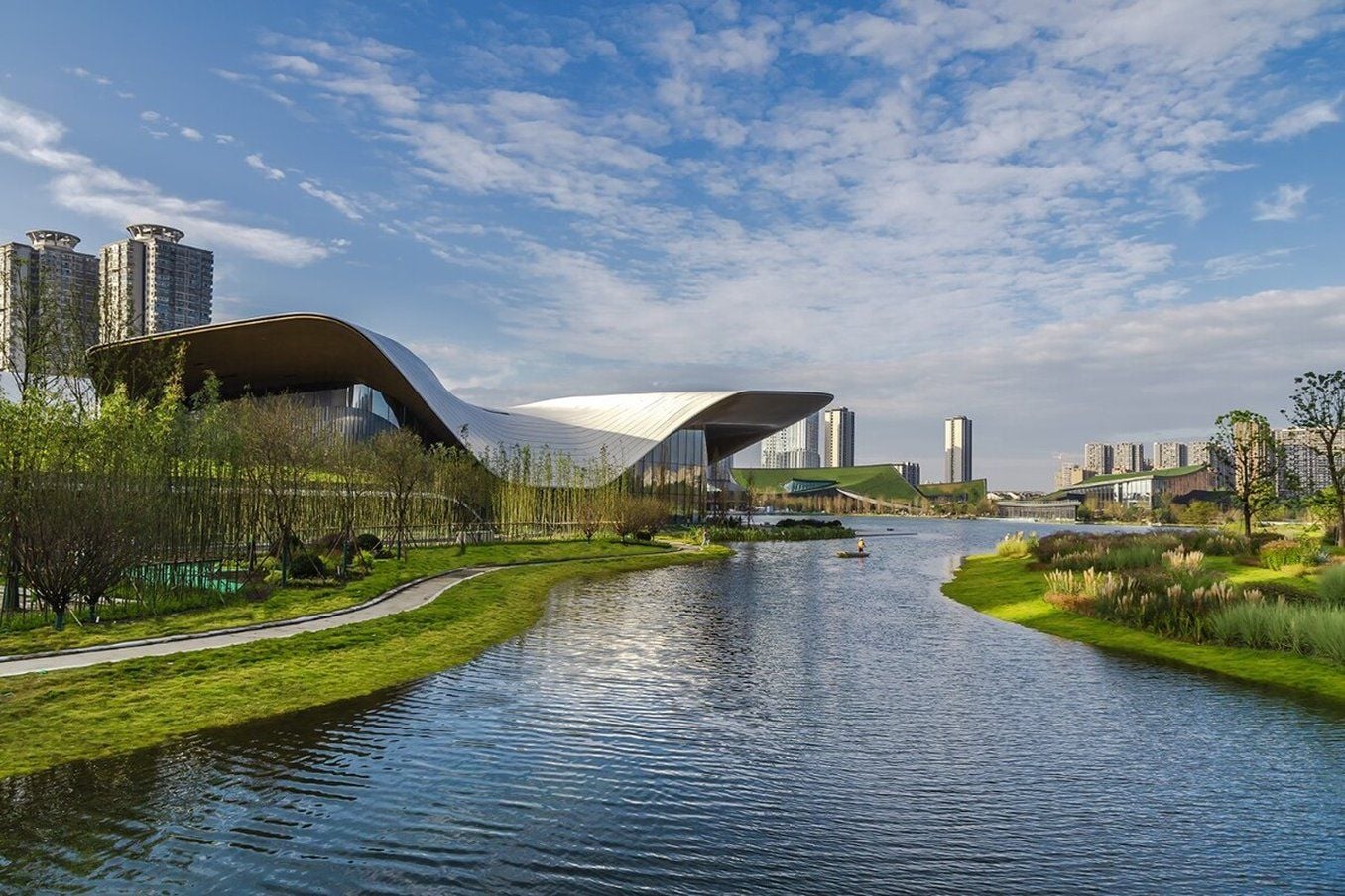 Ein modernes Gebäude mit einem markanten, gewölbten Dach, einem Fluss im Vordergrund und einer Stadtlandschaft im Hintergrund.