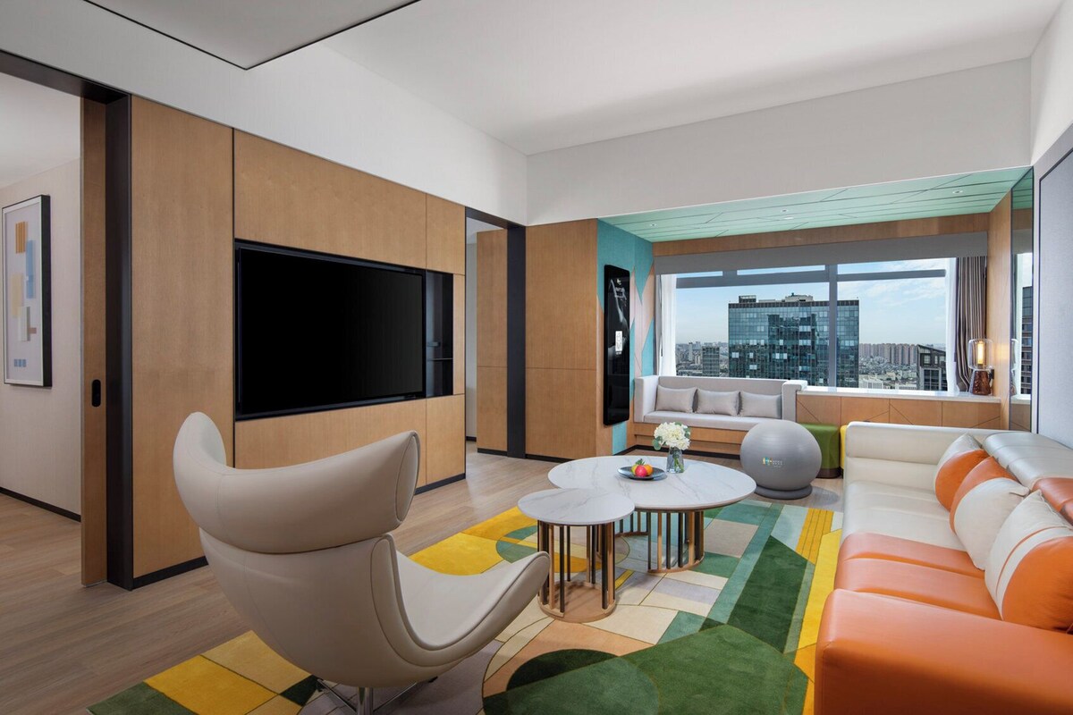 A modern living room with a large flat-screen TV, comfortable seating, a city view, and a large window.