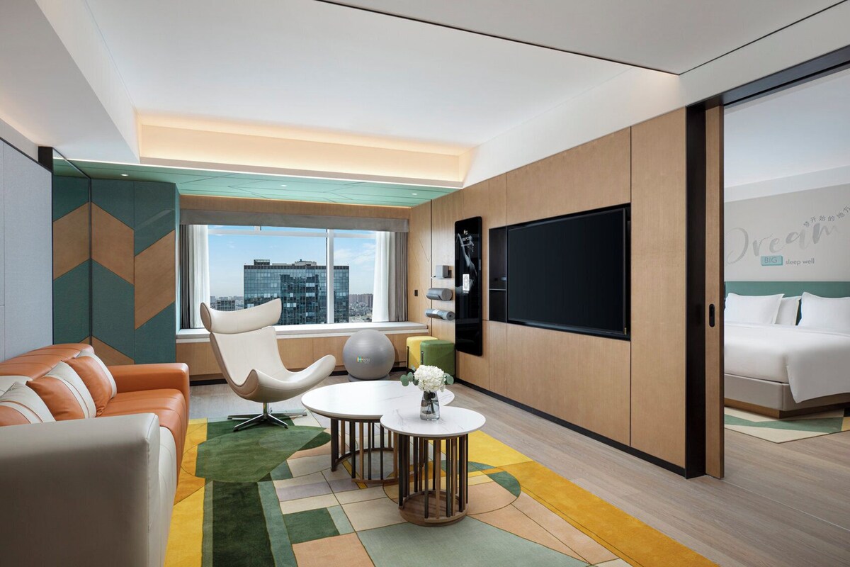 A modern hotel room with a large bed, a seating area with a sofa and chairs, a small table with a vase, and a large window offering a city view.