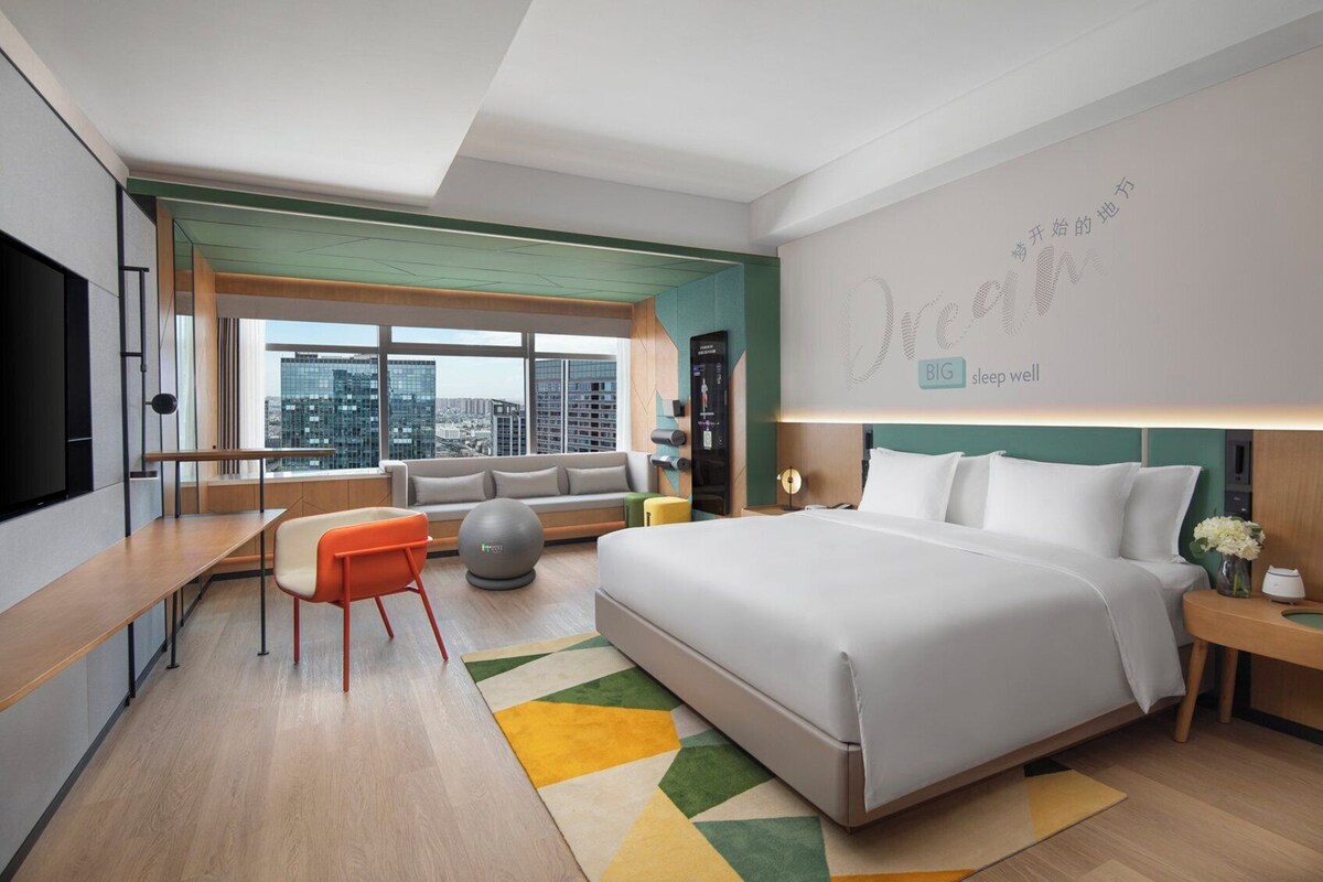 A modern hotel room with a large bed, a sofa, a desk, and a view of the city.
