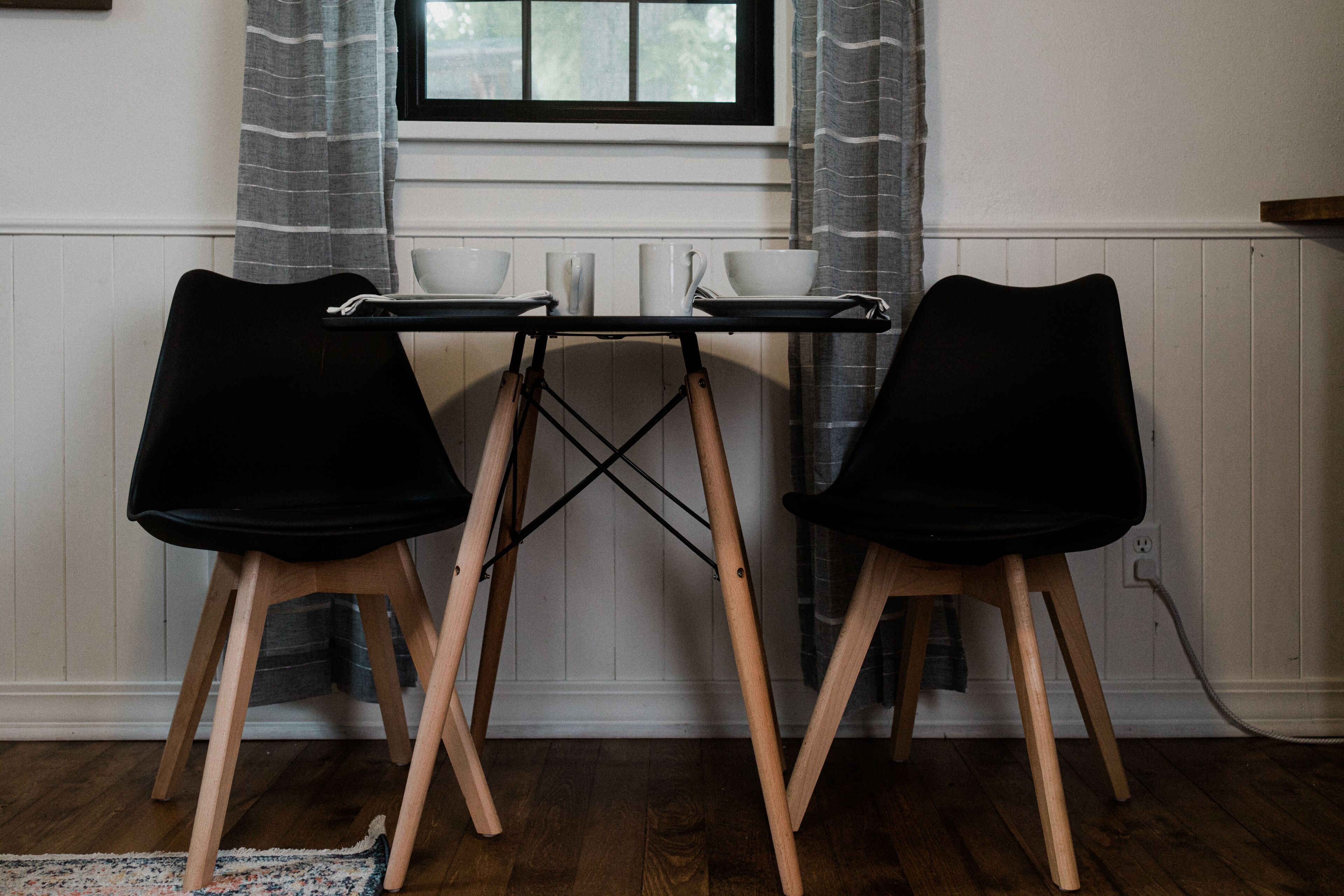 Une pièce avec un plancher en bois, une fenêtre avec des rideaux et deux chaises noires aux pieds en bois.