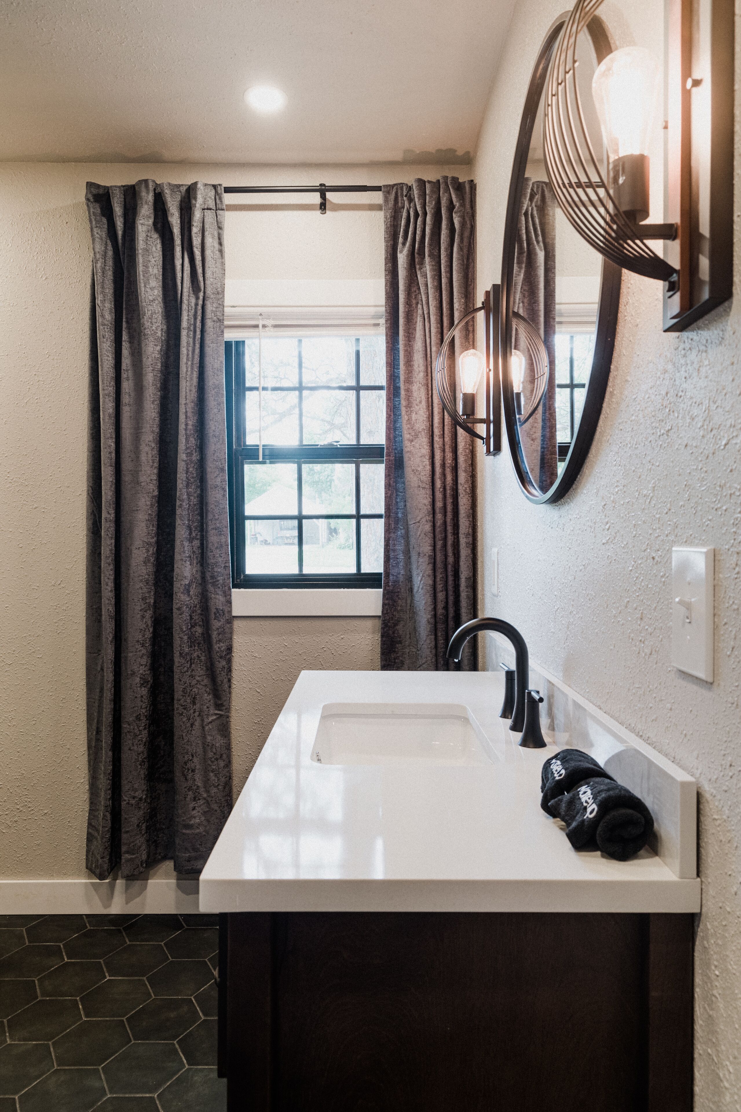 Une salle de bain avec un lavabo blanc, un meuble en bois foncé et une fenêtre avec des rideaux.