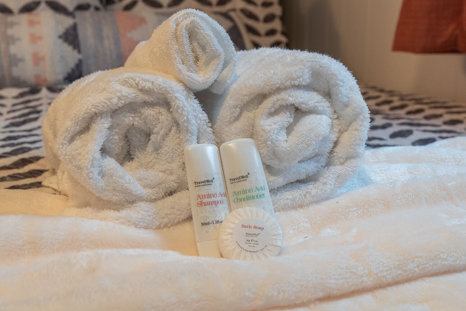A bed with rolled white towels and two TravelWell products: Amino Acid Shampoo and Conditioner.