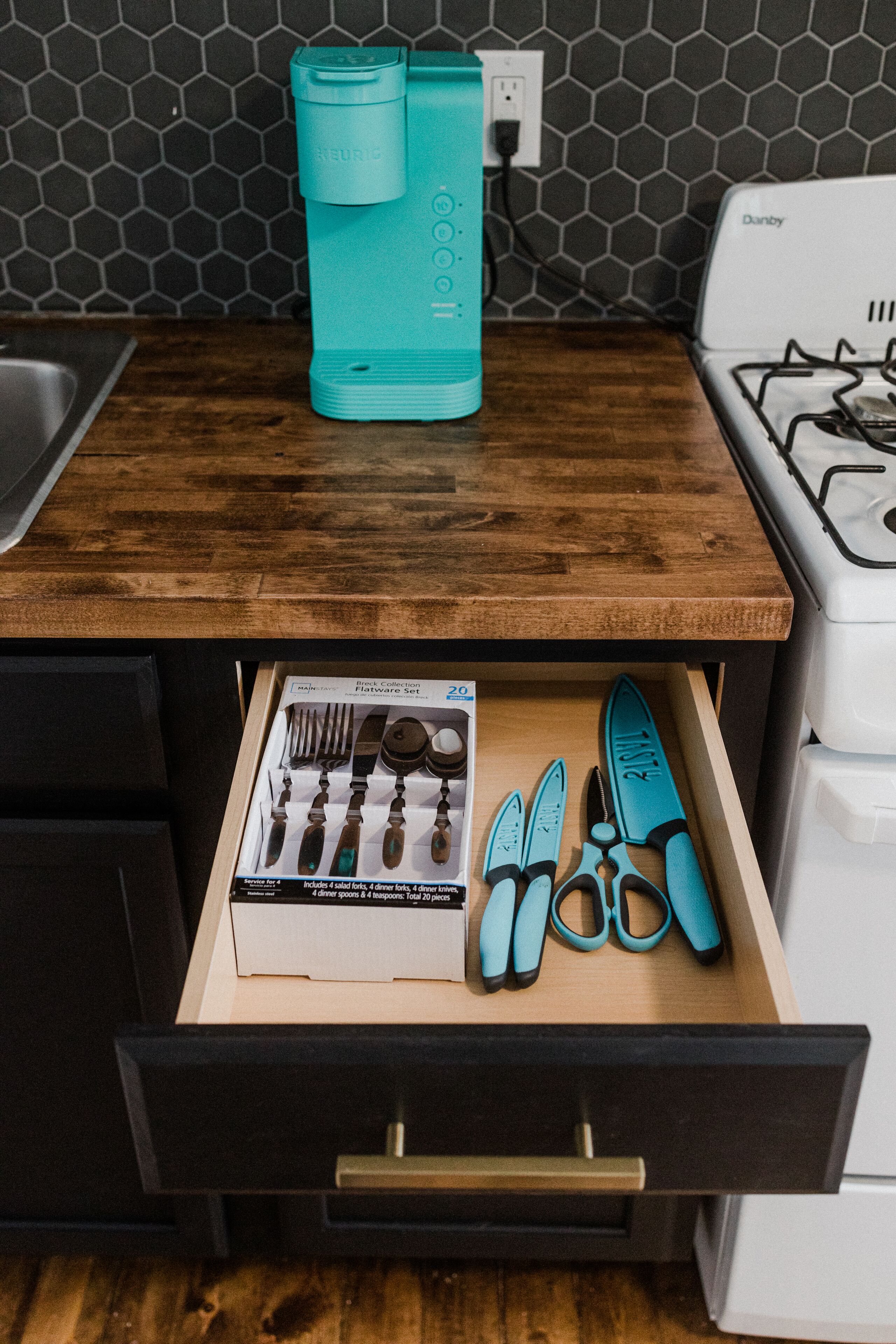 Een keukenblad met een waterfles in teal, een gasfornuis en een lade met bestek en scharen.