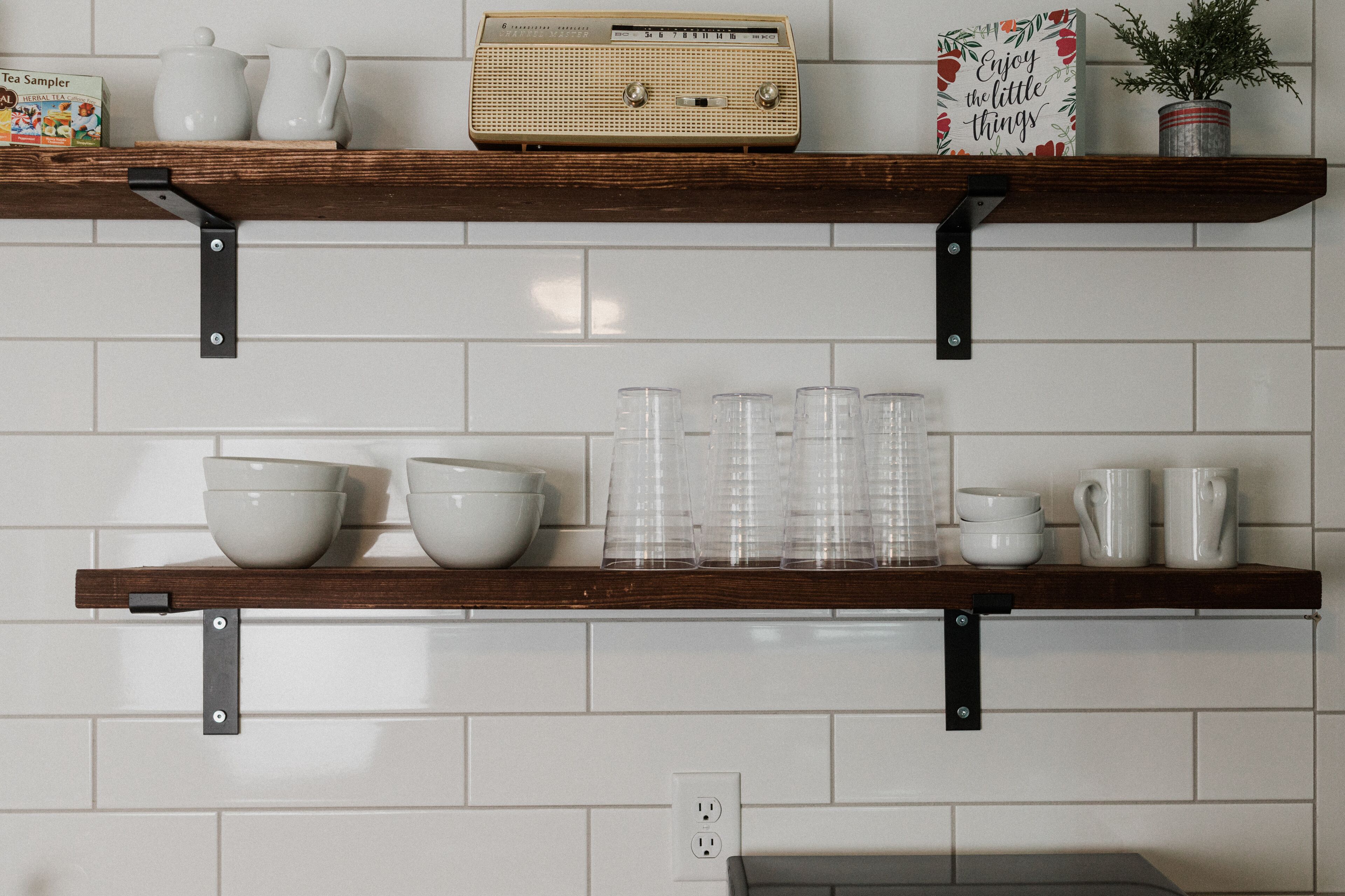 Dos repisas de madera fijadas en una pared de azulejos blancos, con varios objetos de cocina como cuencos, tazas y un radio.