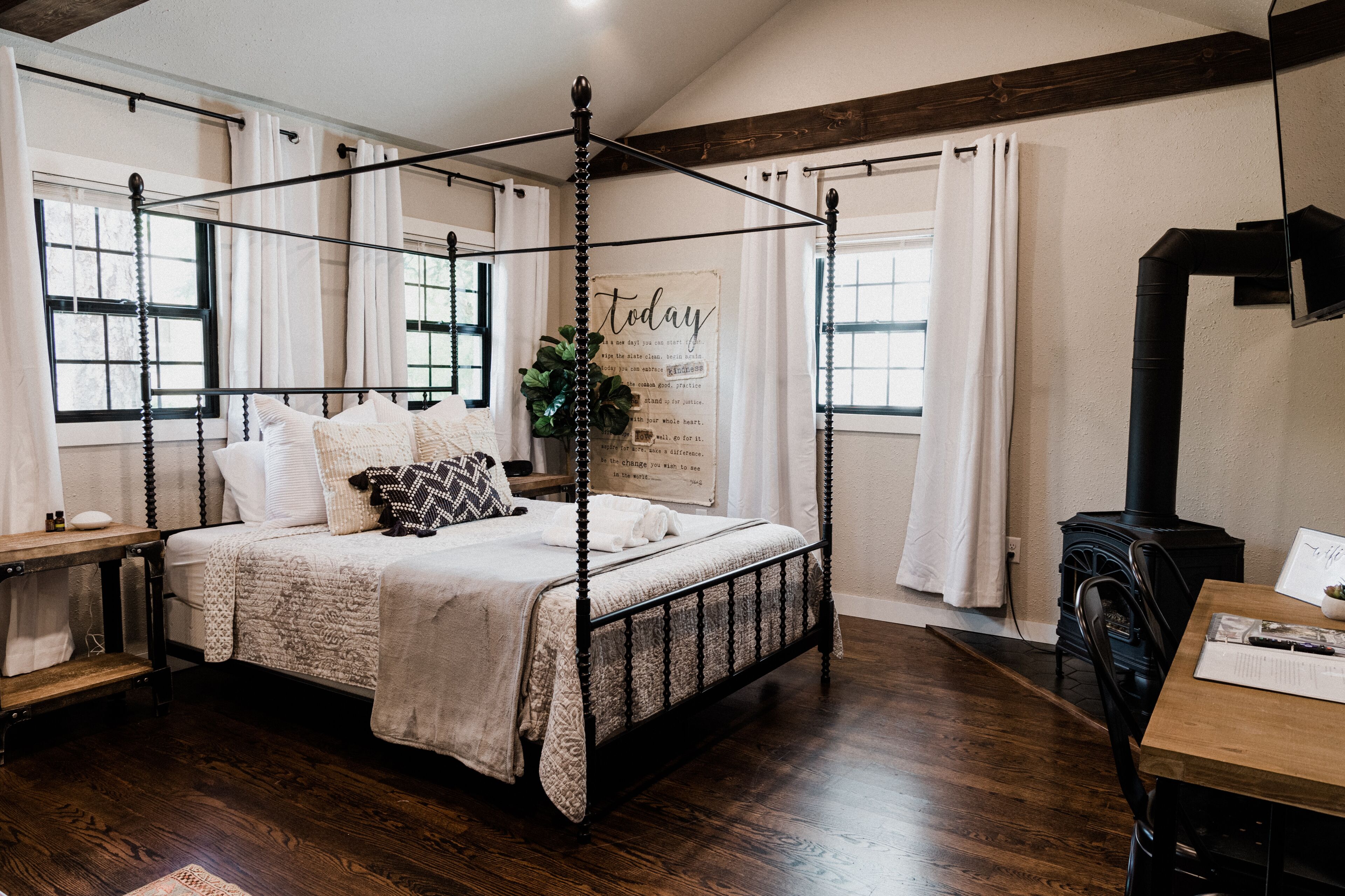 A four-poster bed with a white bedspread and multiple pillows, a wooden nightstand, a black heater, and a wooden table with a chair.