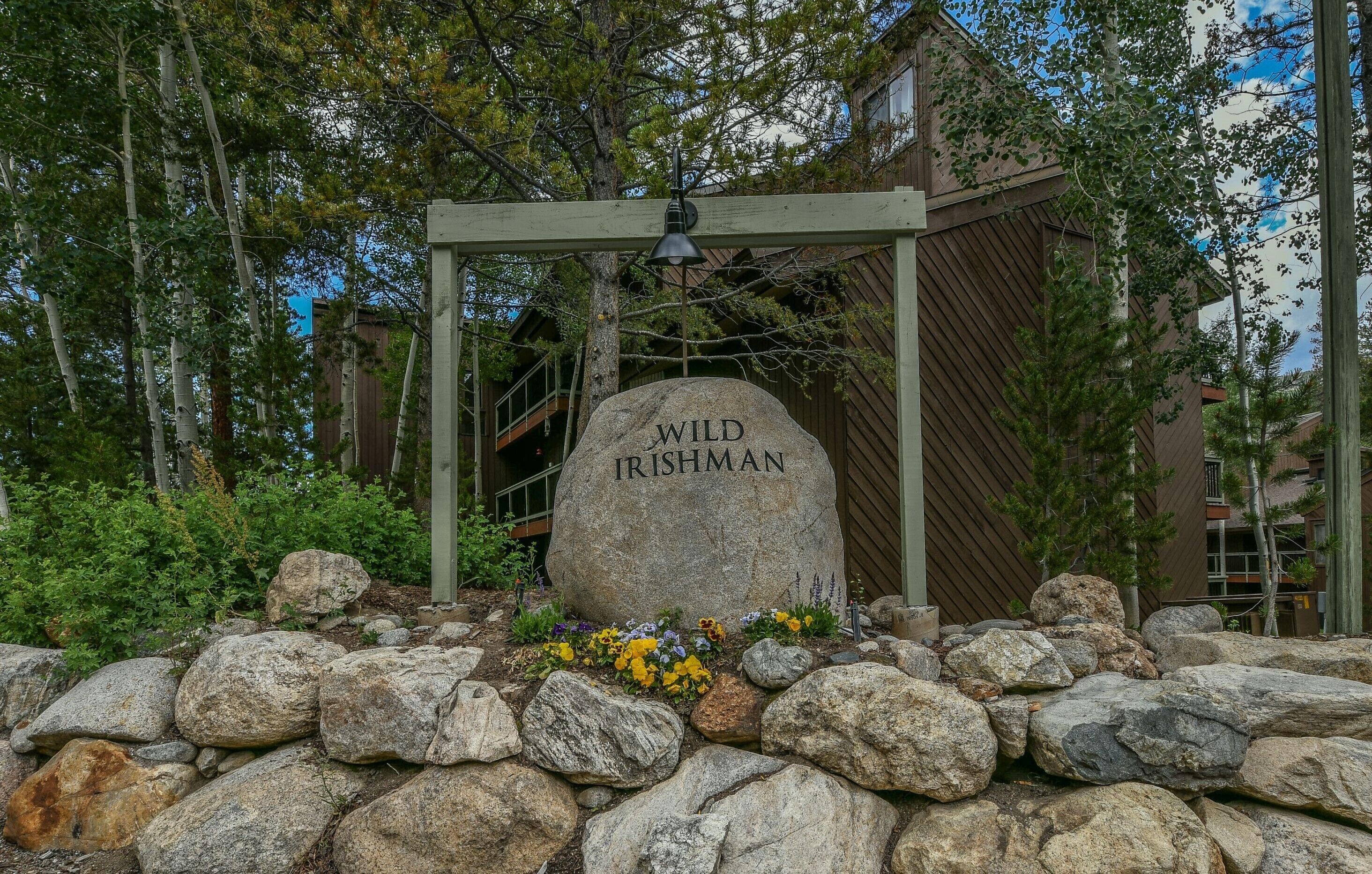 A stone marker with "WILD IRISHMAN" inscribed, surrounded by rocks and flowers, with a wooden pergola overhead and buildings in the background.