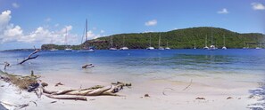 A beach with boats, clear water, and a forested hill in the background.