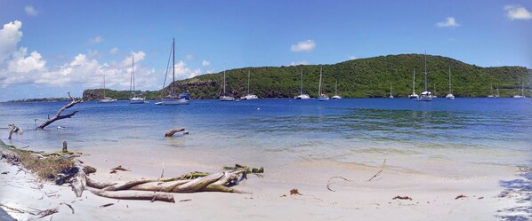 A beach with boats, clear water, and a forested hill in the background.