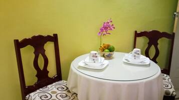 A dining table with two chairs, a vase with flowers, and a bowl of fruit.