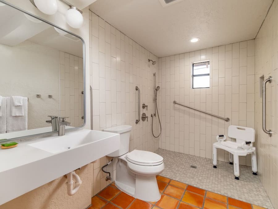 Een badkamer met een wit wastafelmeubel, toilet en douche. Er is een raam en een witte stoel.
