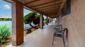 A covered outdoor area with a concrete floor, metal chairs, and a view of a tree and a building in the background. - Pecos Trail Inn (Santa Fe)