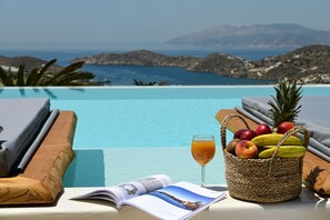 A poolside view with a wicker basket of fruits, a glass of juice, and a magazine.
