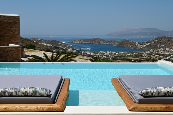 A poolside view with two lounge chairs, a clear blue pool, and a scenic view of a coastal town and mountains.