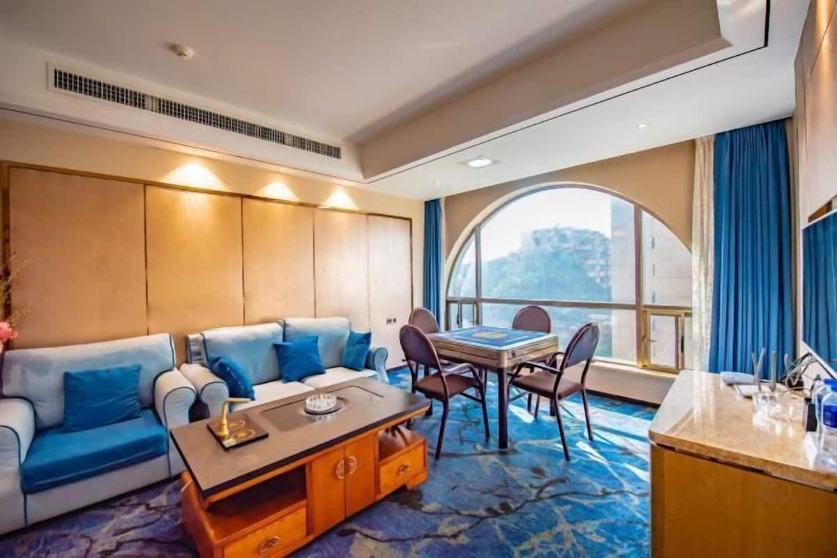 A modern hotel room with a sofa, a dining table with chairs, a small wooden side table, and a large window offering a view.