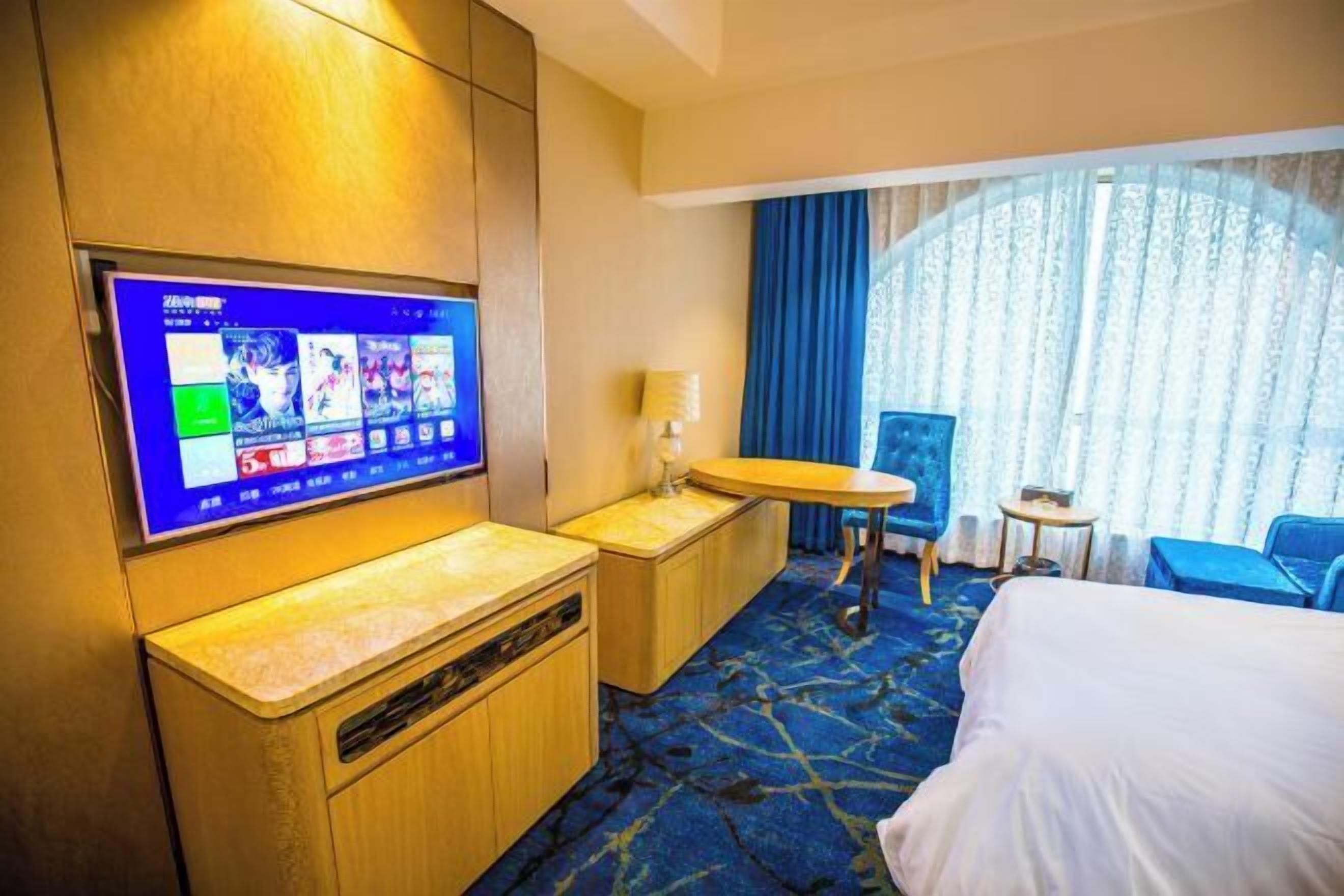 A hotel room with a large flat-screen TV mounted on the wall, a bed, a desk with a chair, a small table, and a window with curtains.
