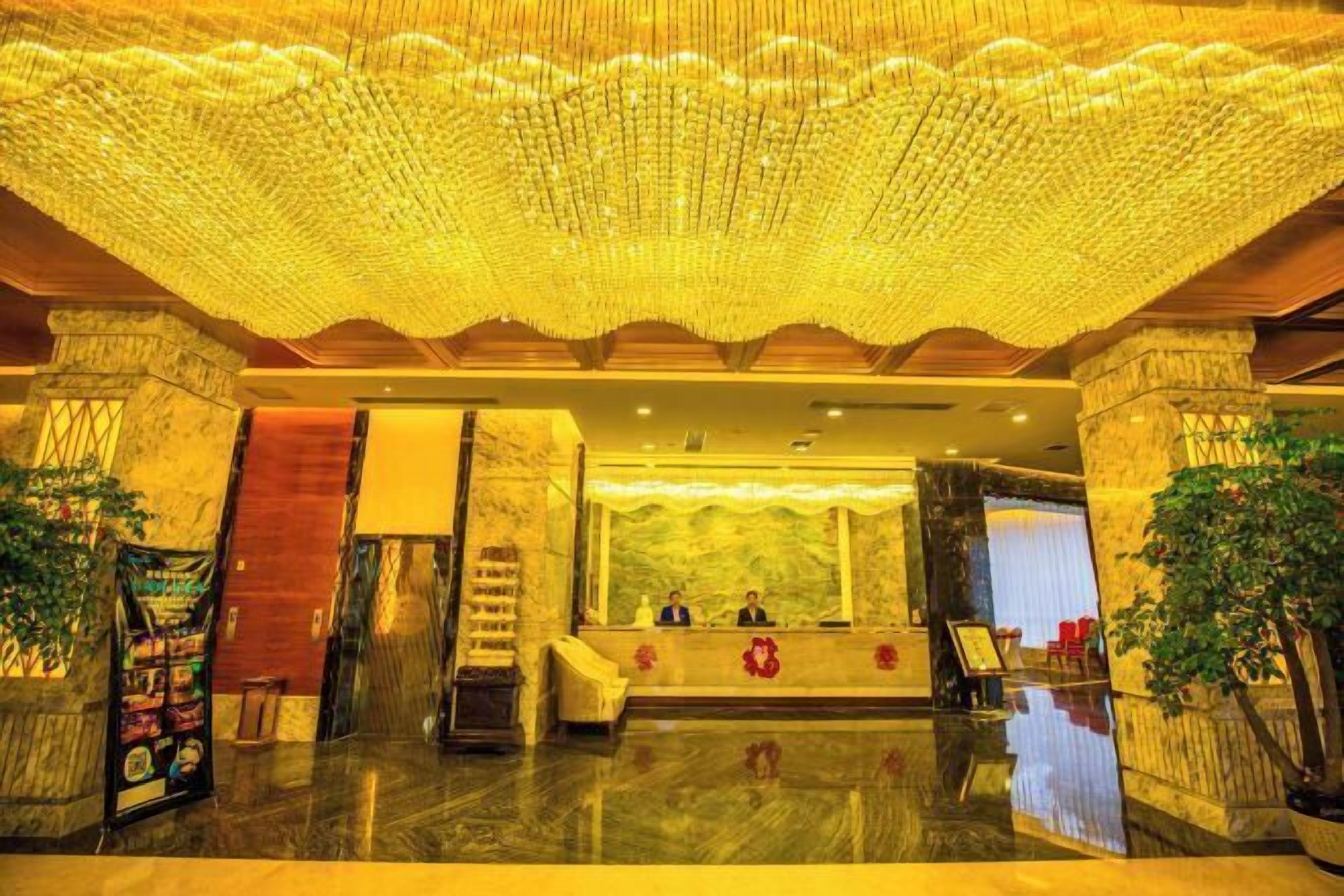 A lobby with a reception desk, two seated individuals, a chandelier, and a large plant.