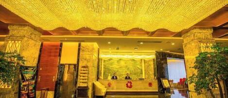 A lobby with a reception desk, two seated individuals, a chandelier, and a large plant.