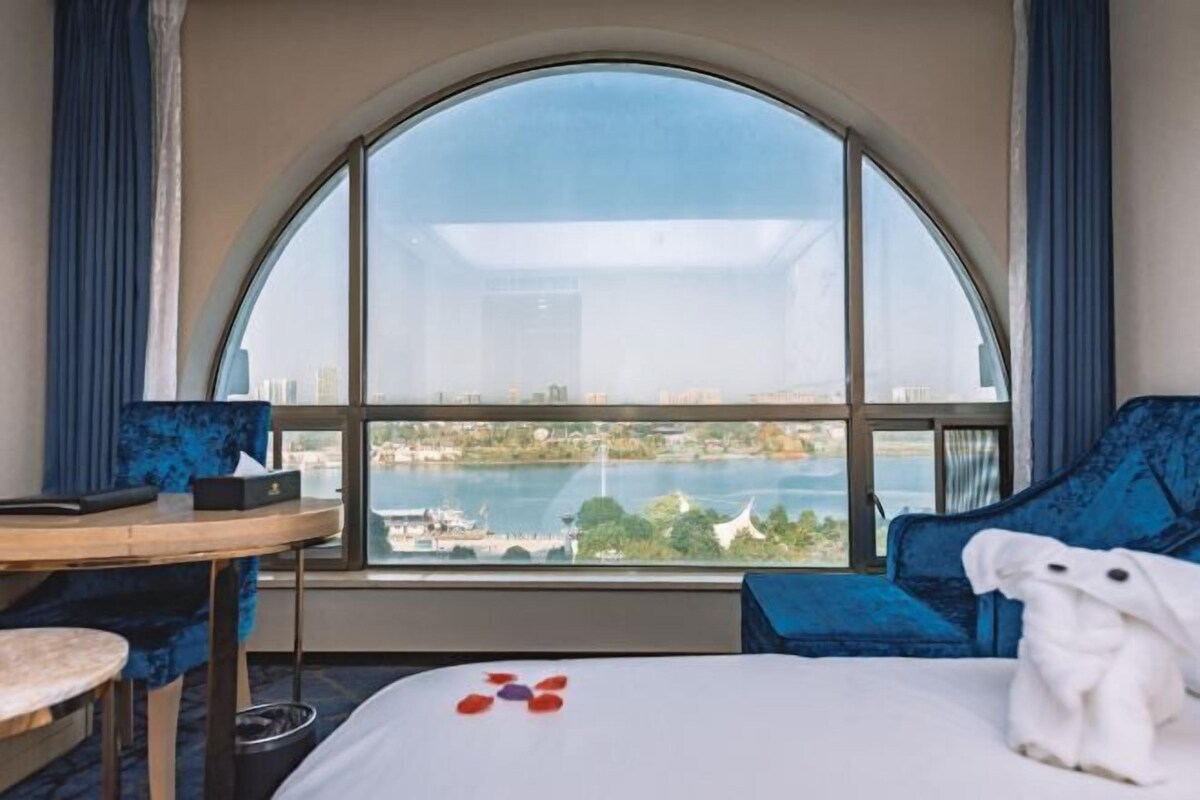 A hotel room with a large arched window, a bed with a white comforter, a blue upholstered chair, a small round table with a black tray, and a white towel on the bed.