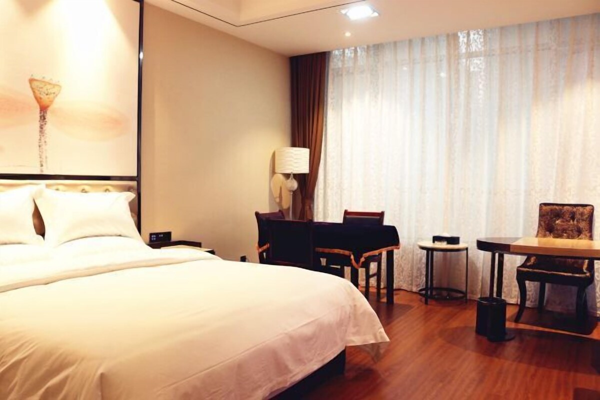 A hotel room with a large bed, a desk, a chair, a lamp, and a view of the outdoors through the window.
