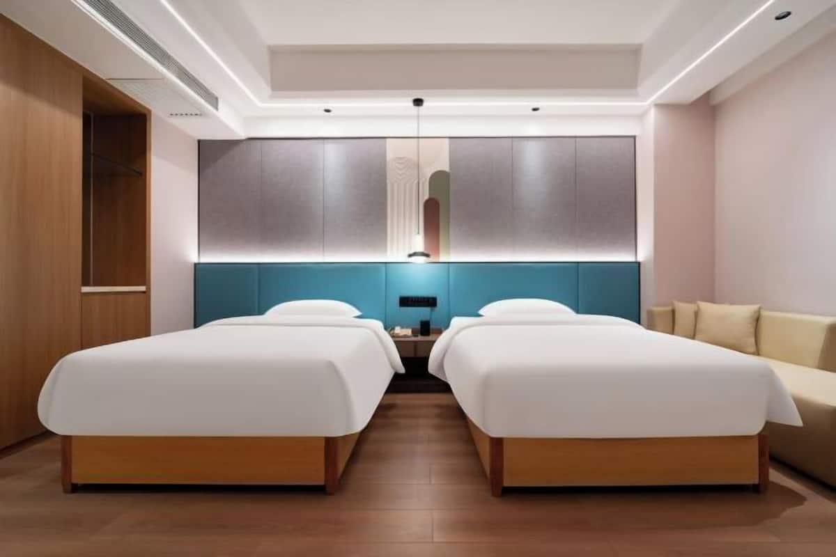 A hotel room with two beds, a nightstand, a wall-mounted lamp, and a wooden wardrobe.