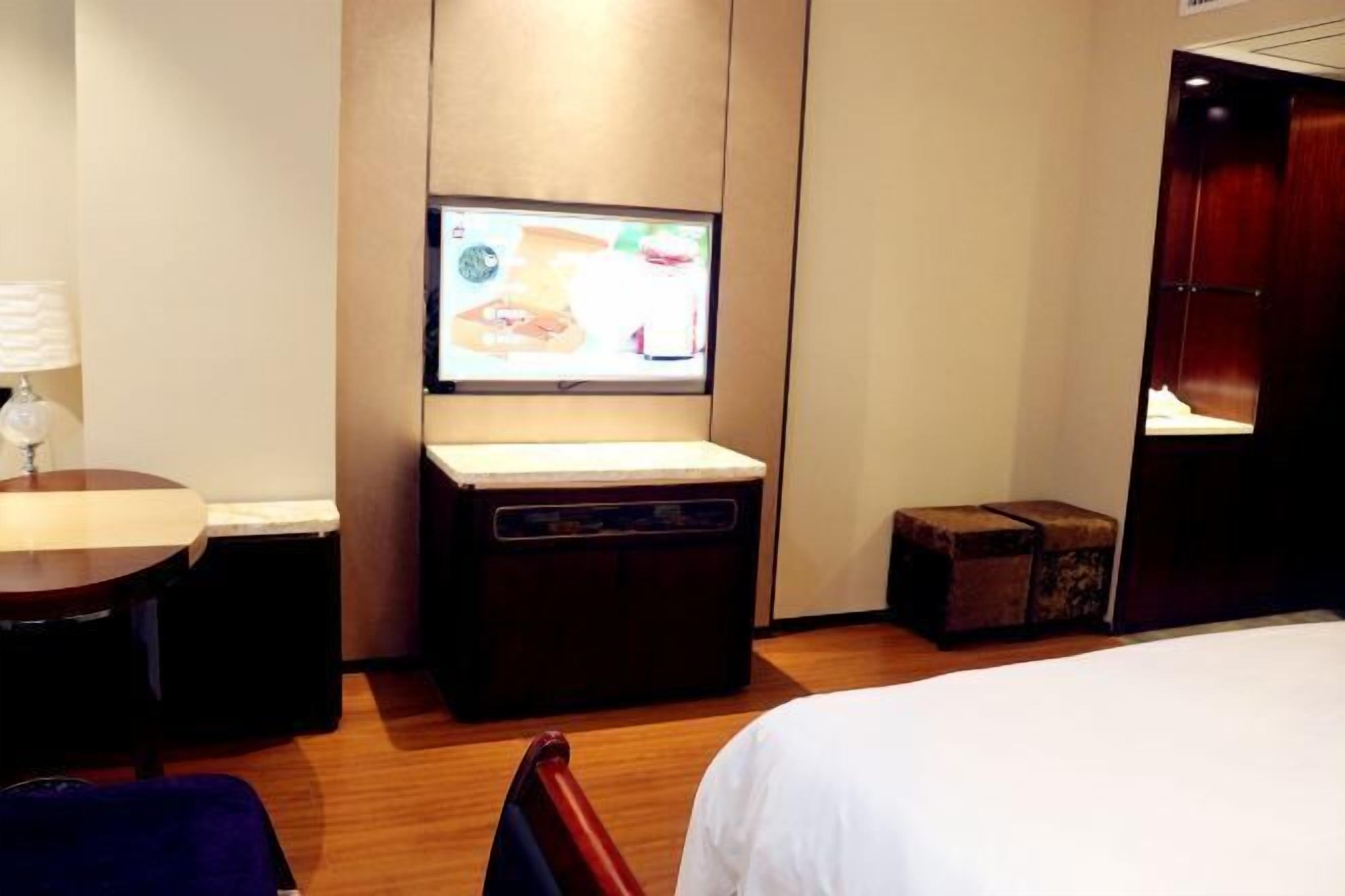 Una habitación de hotel con una cama, televisión montada en la pared, una mesita con lámpara y suelo de madera.
