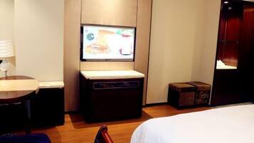 Una habitación de hotel con una cama, televisión montada en la pared, una mesita con lámpara y suelo de madera.