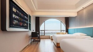 Une chambre d’hôtel dotée d’un grand téléviseur mural, d’un bureau avec une chaise, de deux lits et d’une grande fenêtre donnant sur un paysage agréable.