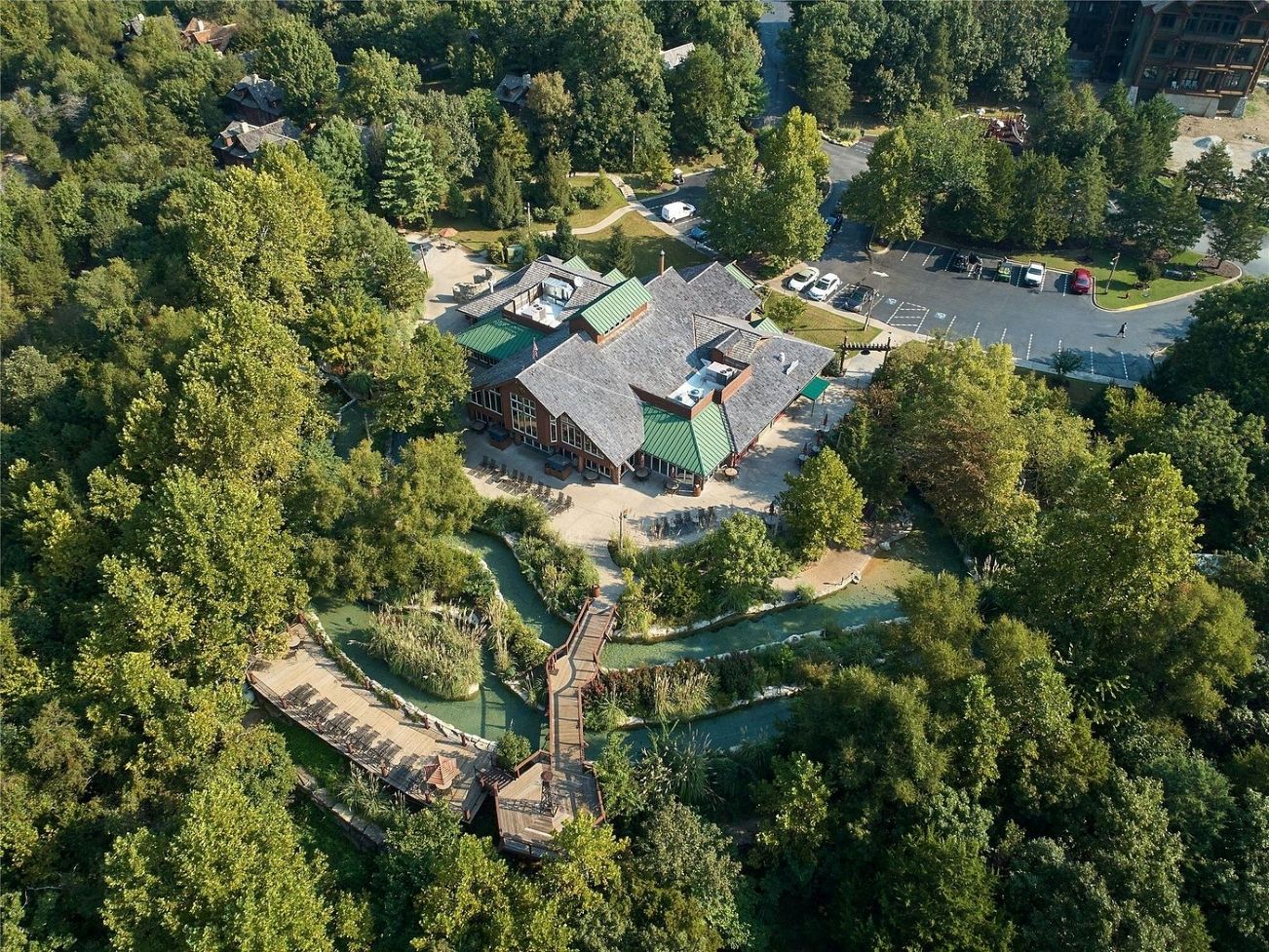 An aerial view of a property with a central building surrounded by dense greenery, a parking area, and a small water feature.