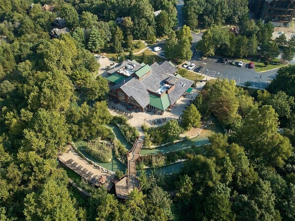Vue aérienne d’une propriété dont le bâtiment principal est entouré d’une végétation luxuriante, avec un espace de stationnement et un petit élément décoratif aquatique.