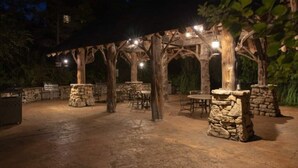 An outdoor patio area with stone pillars, tables, and chairs under a wooden pergola with string lights.
