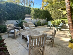 Un patio con mobiliario de madera, incluyendo sillas y una mesa, rodeado de macetas con plantas y un muro de piedra.