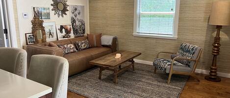 Una sala de estar con un sofá de cuero marrón, una mesa de centro de madera, un tapete estampado y una ventana con persianas.