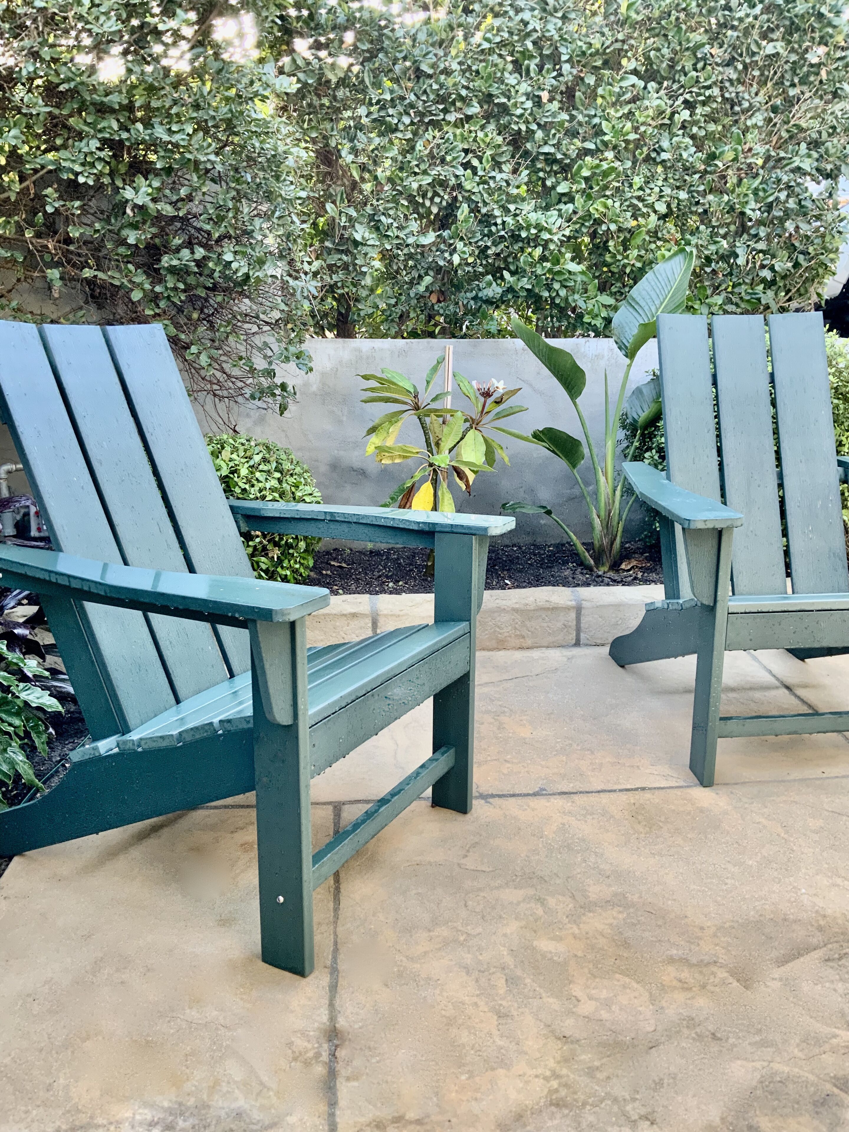 Zwei grüne Adirondack-Stühle mit einem Blumentopf dazwischen auf einer Beton-Terrasse.
