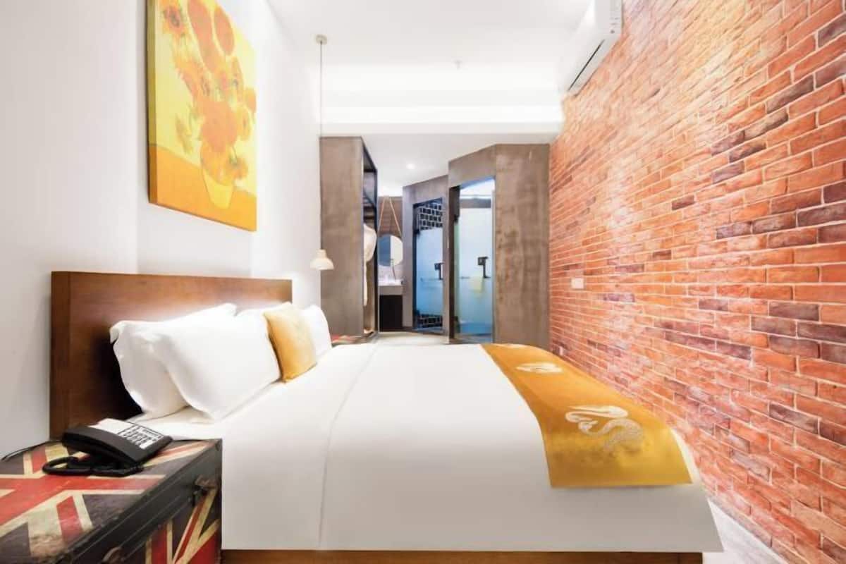 A hotel room with a brick wall, a bed with white bedding, a bedside table with a phone, and a painting on the wall.
