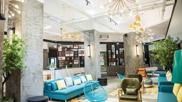 A modern lobby with a blue sofa, a green armchair, and a blue fan. There are bookshelves and a large plant in the background.