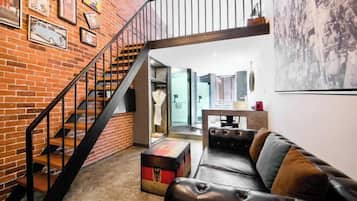 Una sala de estar moderna con pared de ladrillo, un sofá de cuero, una escalera y un cuadro de un paisaje urbano.