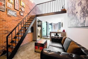 Un salon moderne avec un mur en briques, un canapé en cuir, un escalier et un tableau représentant un paysage urbain.