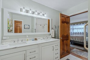 Une salle de bain avec un grand miroir, deux lavabos et une porte en bois donnant sur une chambre.