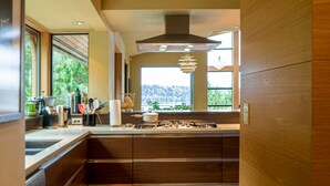 A modern kitchen with wooden cabinets, a built-in stove, and a large window offering a view of greenery. - Modern Hillhouse with Lake Washington Views (Kirkland)