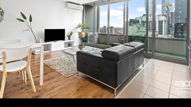 Uma sala de estar moderna com um sofá preto, uma mesa de centro de vidro e uma TV sobre um suporte branco. Há uma área de jantar com uma mesa e cadeiras de madeira. Grandes janelas oferecem vista para o horizonte da cidade.