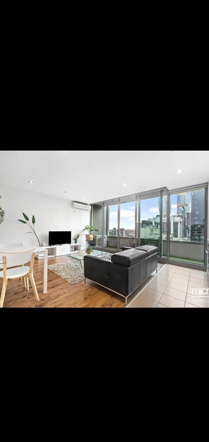 Ein modernes Wohnzimmer mit einer schwarzen Couch, einem Glastisch und einem Fernseher auf einem weißen Lowboard. Es gibt einen Essbereich mit einem Holztisch und Stühlen. Große Fenster bieten einen Blick auf die Skyline der Stadt.