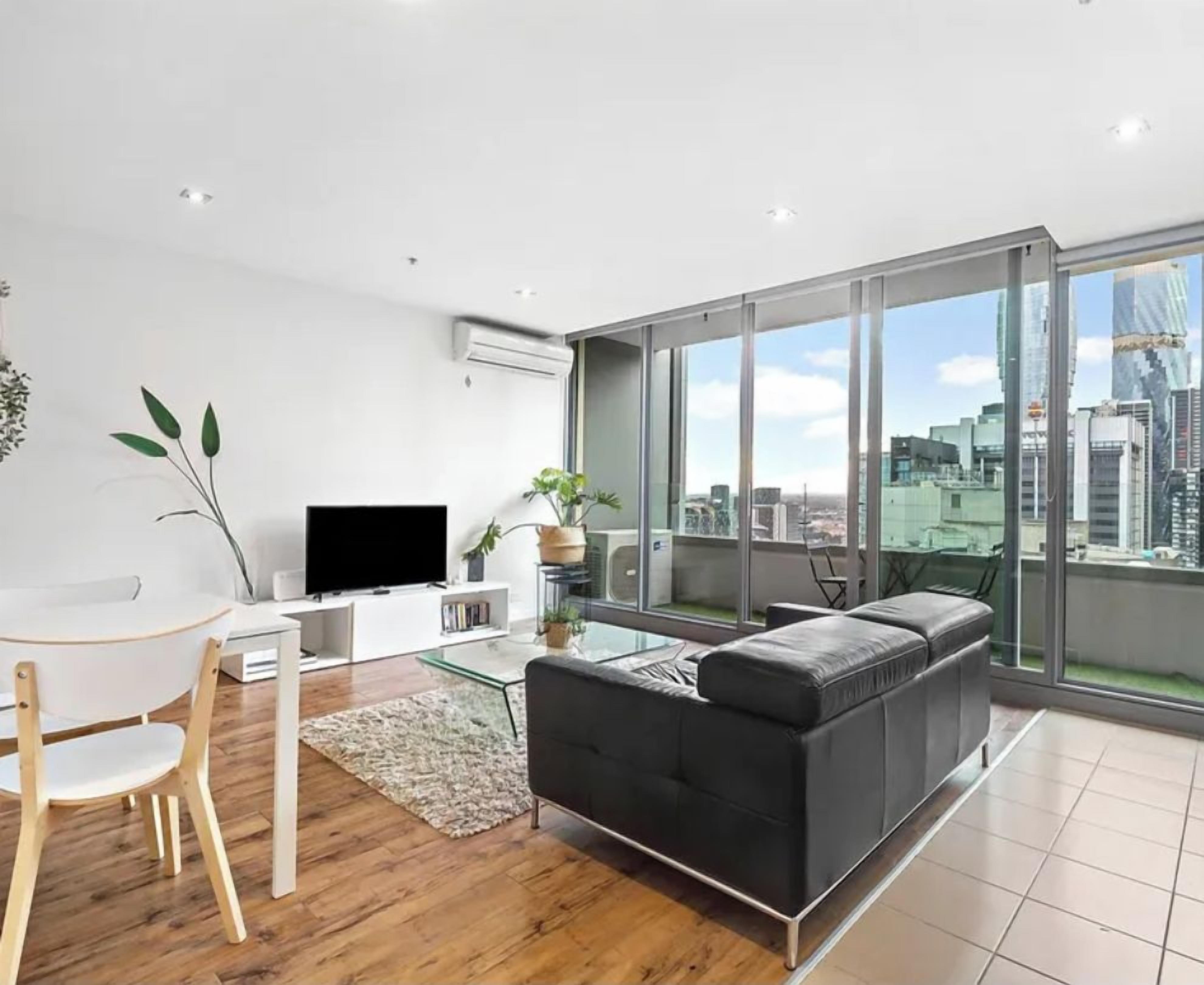 A modern living room with a black sofa, a glass coffee table, and a TV on a white stand. There is a dining area with a wooden table and chairs. Large windows offer a view of the city skyline.
