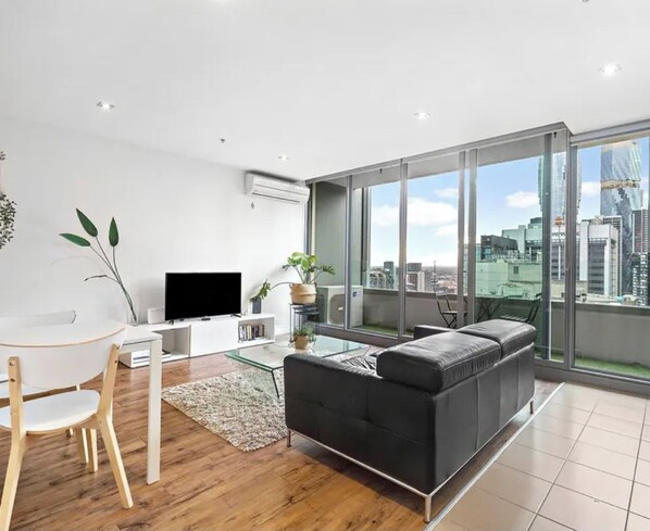 A modern living room with a black sofa, a glass coffee table, and a TV on a white stand. There is a dining area with a wooden table and chairs. Large windows offer a view of the city skyline.