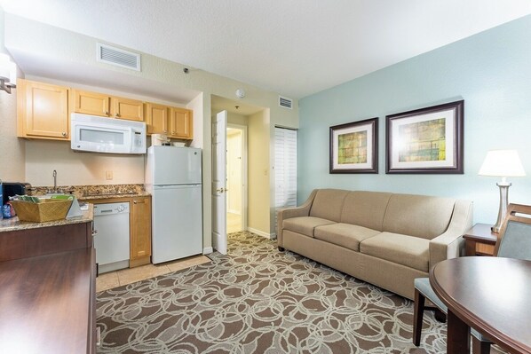 A compact kitchen with a microwave, refrigerator, and dishwasher. There is a beige sofa and a small table with a lamp. Two framed pictures are on the wall.