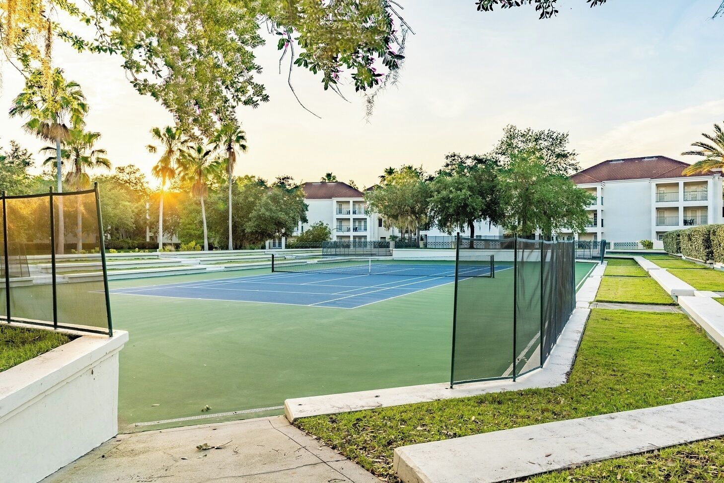 A tennis court with a net, surrounded by a fence, and adjacent to a building with balconies.