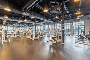 Un gimnasio amplio con diversas máquinas de ejercicio, ventanas grandes y lámparas colgantes.