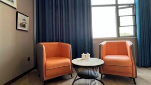 A room with two orange chairs, a small round table with a white top, and a window with blue curtains. - Feng Huang Business Hotel (Luxi)