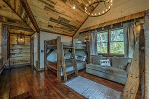 A rustic cabin interior with wooden walls, a loft bed, a sofa, and a large window.
