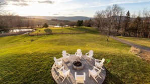 A circular outdoor seating area with white chairs around a fire pit, set in a grassy field with a lake and trees in the background. - Tranquil Lodge with A+ Amenities (Naples)